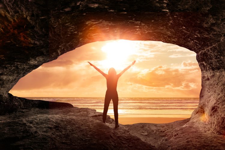 A woman watches the sunrise from a cave by the sea